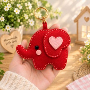 May include: A red felt elephant keychain featuring a light pink heart appliqué on its ear, a small light pink circular cheek, and a black bead eye. The elephant is attached to a gold-toned metal split ring by a small red fabric loop.