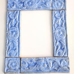 Op de afbeelding: Een vierkant frame gemaakt van acht lichtblauwe keramische tegels. Elke tegel heeft een verhoogd bloemen- en krulwerkpatroon. Het frame omringt een witte ruimte in het midden. De tegels zijn rechthoekig.