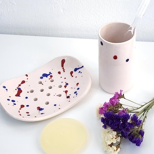 Op de afbeelding: Een lichtroze keramische zeepbakje met blauwe en rode verfspatten, een bijpassende tandenborstelhouder en een stuk zeep. Een takje paarse en witte bloemen staat naast de houder.