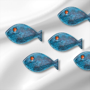 Puede incluir: Varios objetos decorativos azules con forma de pez y ojos naranjas están dispuestos sobre un fondo blanco. Los peces tienen una superficie texturizada con detalles en azul oscuro y morado. Los peces miran en diferentes direcciones.
