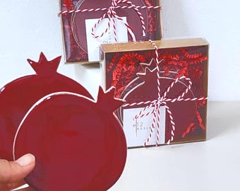 Può includere: Sottobicchieri rossi a forma di melograno con bordi bianchi, presentati in confezioni regalo con carta crespa rossa e legati con spago rosso e bianco. Le scatole hanno un coperchio trasparente e un'etichetta con "e12 design".