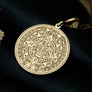 May include: A gold-toned circular pendant featuring an intricate Aztec calendar design with detailed engravings. The pendant is attached to a thin matching metal chain, showcasing a polished finish and a complex, symmetrical pattern across its surface.