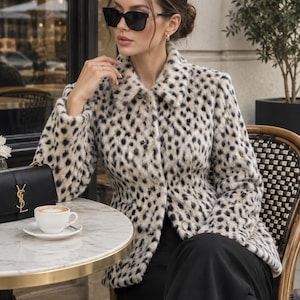 May include: A long-sleeved, button-down jacket featuring a cream-colored faux fur texture with a black leopard print pattern, a standard collar, and a tailored fit. The jacket is worn with black trousers, black sunglasses, and a black handbag with a gold logo.