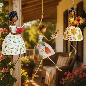 Puede incluir: Tres figuras colgantes con personajes estilizados de cabello oscuro. Uno lee un libro rosa, otro sostiene un pincel y una paleta, y el tercero lleva una calabaza y hojas de otoño. Cada uno viste un vestido acampanado con motivos florales o de puntos.