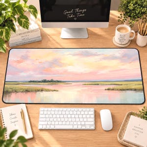 May include: A rectangular desk mat featuring a printed landscape illustration of a marshland at sunset with soft pink, yellow, and blue hues reflecting on the water. The mat has a smooth surface and black edges, positioned on a wooden desk.