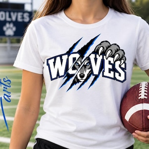 Puede incluir: Una camiseta blanca de manga corta con un gráfico de la palabra WOLVES en letras azules y blancas. El diseño incluye una cara de lobo asomándose a través de marcas de garras y una garra gris sobre la parte superior del texto.