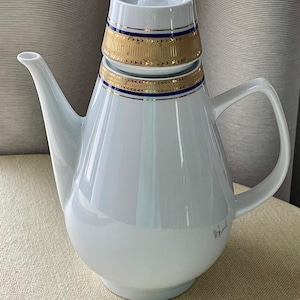Op de afbeelding: Een witte porseleinen theepot met een taps toelopend lichaam, een gebogen handvat en een lange tuit. Het deksel en de bovenhals van de theepot zijn versierd met een brede gouden band, een dunne donkerblauwe streep en een geometrisch goudpatroon.