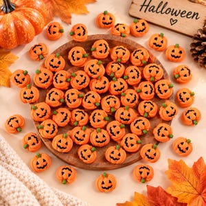 Può includere: Una collezione di piccole perline arancioni a forma di zucca con volti di jack-o'-lantern dipinti in nero. Le perline sono sparse sopra e intorno a un vassoio circolare in legno, accompagnate da un cartello in legno con il testo Halloween.