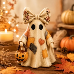 Pode incluir: Uma estatueta de fantasma cor de creme com um laço grande na cabeça decorado com pequenas rosas cor-de-rosa. O fantasma tem um padrão de retalhos com quadrados laranja, amarelos e cinzentos e um coração, e segura um pequeno balde de abóbora.