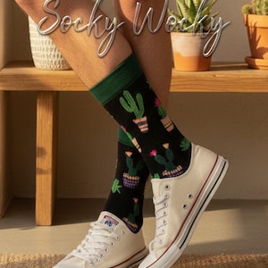 Op de afbeelding: Zwarte sokken met een herhalend patroon van cactussen in potten met roze en groene accenten, afgewerkt met een donkergroene geribbelde boord. De sokken worden gedragen met witte canvas sneakers. De tekst "Socky Wocky" staat in het wit.