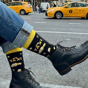 Puede incluir: Calcetines negros de caña media con un patrón repetitivo de taxis amarillos de Nueva York y el texto NYC TAXI en letras doradas. Los calcetines tienen un puño amarillo sólido en la parte superior y se usan con jeans azul oscuro y botas negras.