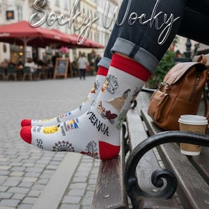 Könnte beinhalten: Hellgraue Crew-Socken mit roten Bündchen und Zehenspitzen, verziert mit bunten Illustrationen von Wiener Sehenswürdigkeiten wie einem Riesenrad, Noten und dem schwarzen Schriftzug "VIENNA". Getragen zu einer dunkelblauen Jeans.