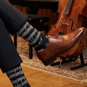 Peut inclure: Une paire de chaussettes noires mi-mollet ornées d'un motif de portée musicale et de notes blanches, portées avec un pantalon sombre et des chaussures de ville à lacets en cuir marron. Les chaussettes présentent une notation musicale détaillée.