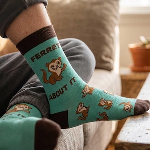 May include: A pair of light teal crew socks featuring a repeating pattern of brown ferrets and the text "FERRET ABOUT IT" in dark brown lettering. The socks have a solid dark brown cuff at the top and a matching dark brown toe section.