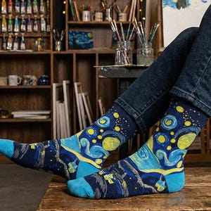 Pode incluir: Um par de meias de cano médio com base azul-escura e um padrão vibrante de redemoinhos em amarelo e azul-claro inspirado na obra A Noite Estrelada. O design inclui corpos celestes amarelos circulares e texturas de pinceladas visíveis.