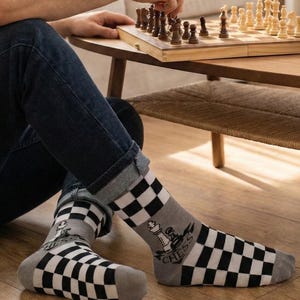 Könnte beinhalten: Ein Paar Socken mit schwarz-weißem Karomuster an Fuß und Bündchen sowie einem grauen Mittelteil, das eine weiße Schachkönig-Grafik und das Wort CHESS in schwarzen Buchstaben auf einem Banner-Design zeigt.