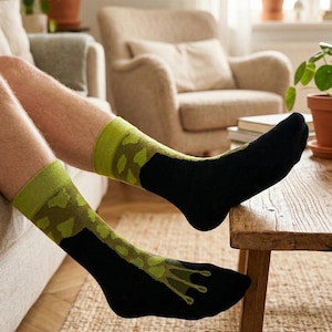 Puede incluir: Un par de calcetines con base de color negro y un patrón inspirado en una rana. El diseño incluye un estampado verde texturizado en la parte superior y un gráfico estilizado de una pata de rana verde que se extiende hacia la zona de los dedos.