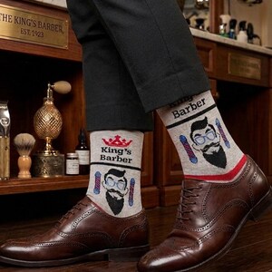 Könnte beinhalten: Hellgraue Socken mit einer Grafik einer bärtigen Person mit blauer Brille, einer roten Krone und Barber-Stangen. Der Text "King's Barber" ist auf die Socken gedruckt. Die Socken werden zu einer schwarzen Hose und braunen Lederschuhen getragen.