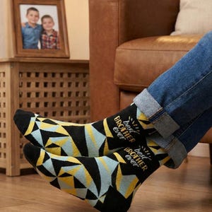 Puede incluir: Un par de calcetines negros con un patrón geométrico de triángulos azul claro y amarillos. Los calcetines muestran el texto "best BROTHER ever" en una fuente blanca y amarilla en la parte superior, con punteras y talones negros.