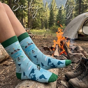 Könnte beinhalten: Hellblaue Socken mit dunkelgrünen Bündchen, Fersen und Zehenbereichen. Die Socken zeigen ein aufgedrucktes Muster aus Bergen, einem Lagerfeuer, dem Schriftzug "Adventure Awaits" in Blau, einer dunkelblauen Landkarte und kleinen weißen Wolken.