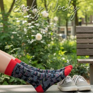 Puede incluir: Un par de calcetines con fondo gris oscuro y un patrón repetitivo de pequeñas caras de gatos negros con ojos amarillos y detalles rojos. Los calcetines tienen puños, talones y punteras de color rojo sólido. El texto "Socky Wocky" aparece arriba.