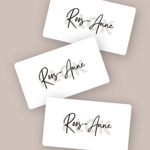 Op de afbeelding: Drie rechthoekige witte kaarten gerangschikt in een overlappend patroon. Elke kaart is voorzien van de naam Roos-Anne in een zwart cursief lettertype. De kaarten hebben afgeronde hoeken en een strak, wit oppervlak met een eenvoudig ontwerp.