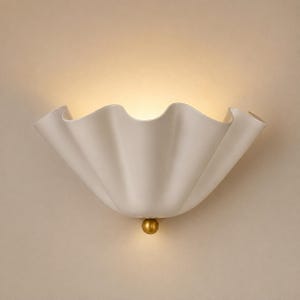 May include: A wall sconce featuring a wide, off-white, ruffled shade with a curved, shell-like silhouette. A small, spherical, metallic gold-toned accent piece is positioned at the bottom centre of the fixture, which emits a warm, soft glow.