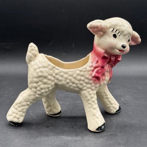 May include: A cream-colored ceramic lamb planter featuring a textured wool finish, a glossy pink bow around its neck, and black painted hooves. The lamb has a hollow opening on its back for plants and a detailed face with dark eyes and a small nose.