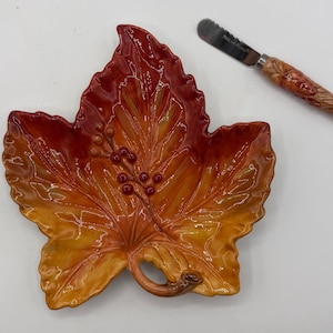 May include: A ceramic leaf-shaped dish featuring a gradient of red, orange, and yellow colors with a raised berry branch design in the center. Beside the dish is a metal spreader knife with a textured, carved wooden handle featuring leaf details.