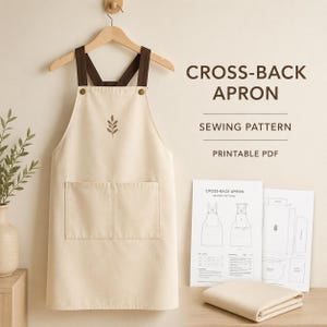 Puede incluir: Un delantal de espalda cruzada color crema con tirantes marrón oscuro y un pequeño bordado de hoja, colgado en una percha de madera. Junto a él hay hojas de patrón de costura impresas con el texto CROSS-BACK APRON SEWING PATTERN PRINTABLE PDF.