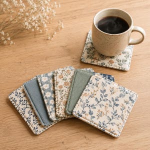 Könnte beinhalten: Ein Set aus quadratischen Stoffuntersetzern mit verschiedenen Blumen- und Unimustern in Blau-, Grün- und Cremetönen. Ein Untersetzer trägt eine gesprenkelte Keramiktasse mit schwarzem Kaffee, während die anderen darunter fächerförmig ausgebreitet sind.