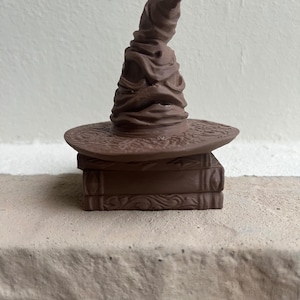 May include: A brown, monochromatic figurine featuring a pointed, creased hat with a wide brim resting atop a stack of two rectangular books. The object has a matte finish and displays detailed textures on the hat's folds and the book covers.