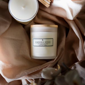 May include: Two frosted glass candles with light-colored wooden lids. One candle is upright with a label reading Embers Edge, Hand Poured Soy Candle, White Oak & Vanilla, 4 oz. The other candle is open with the lid placed beside it.