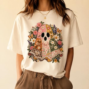 Peut inclure: Un t-shirt à manches courtes blanc cassé avec un motif central représentant un fantôme composé de divers tissus en patchwork, incluant des motifs vichy, floraux et à pois, entouré d'un arrangement coloré de fleurs style broderie.