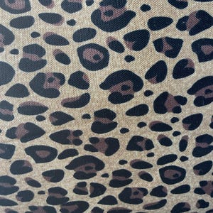 May include: A woven fabric featuring a repeating leopard print pattern. The design consists of dark brown and black irregular spots set against a light tan, textured background, showcasing a clear weave structure throughout the material surface.