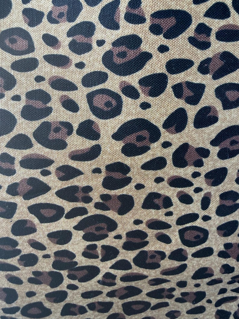 May include: A woven fabric featuring a repeating leopard print pattern. The design consists of dark brown and black irregular spots set against a light tan, textured background, showcasing a clear weave structure throughout the material surface.