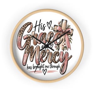 Könnte beinhalten: Eine runde Wanduhr mit hellem Holzrahmen und schwarzen Zeigern. Das Zifferblatt hat einen weißen Hintergrund mit dem Text His Grace and Mercy has brought me through in rosa und Leopardenmuster-Schrift, einem Holzkreuz mit rosa Schleife und Herzen.
