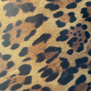 May include: A close-up view of a fabric featuring a classic leopard print pattern. The design consists of irregular black and dark brown spots scattered across a golden tan background, creating a repeating animal-inspired motif on the textile surface.