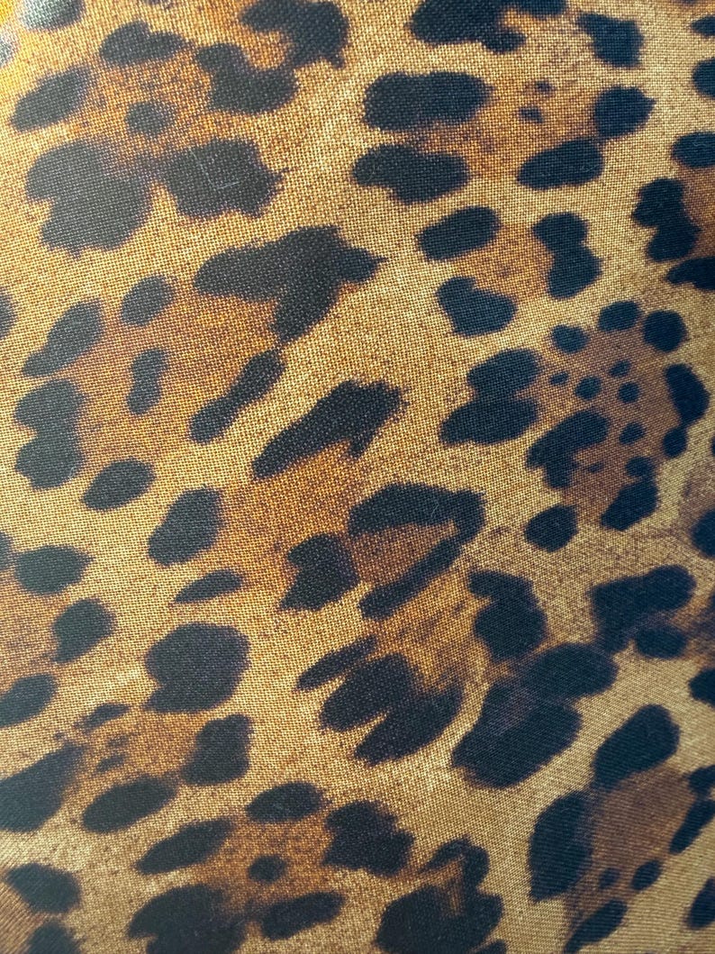 May include: A close-up view of a fabric featuring a classic leopard print pattern. The design consists of irregular black and dark brown spots scattered across a golden tan background, creating a repeating animal-inspired motif on the textile surface.