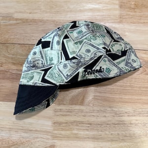 May include: A welding cap featuring a repeating pattern of United States dollar bills in shades of green, black, and white. The cap has a solid black brim and includes the word "Bornado" embroidered in black thread on the side panel.