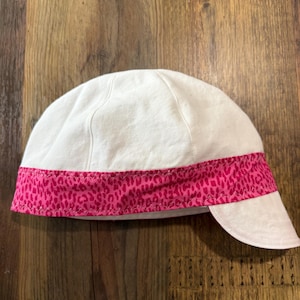May include: A white cap featuring a horizontal band of bright pink fabric with a dark pink leopard print pattern. The cap has a rounded crown constructed from multiple panels and a short, curved brim extending from the front lower edge.