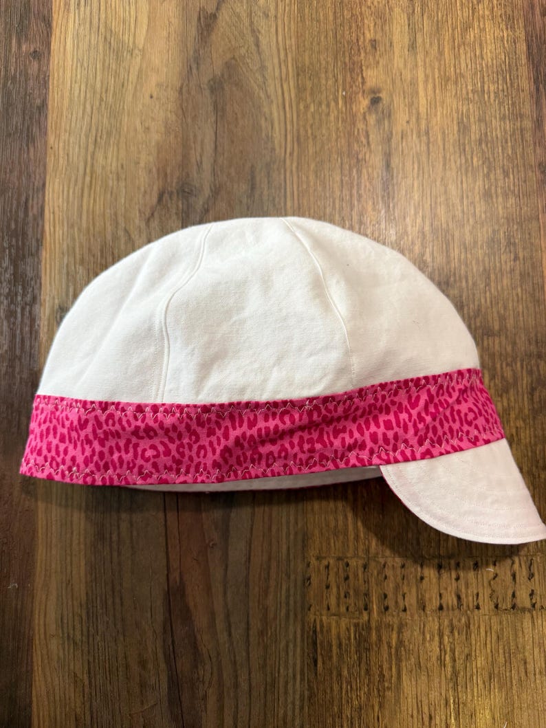 May include: A white cap featuring a horizontal band of bright pink fabric with a dark pink leopard print pattern. The cap has a rounded crown constructed from multiple panels and a short, curved brim extending from the front lower edge.