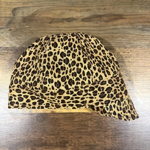 May include: A welding cap featuring a tan and brown leopard print pattern. The cap is constructed with multiple fabric panels joined by visible stitching, creating a rounded crown shape with a short, curved brim extending from the front.
