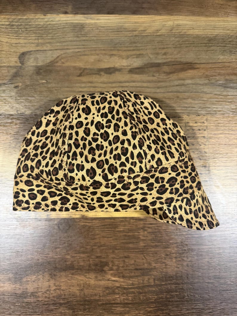 May include: A welding cap featuring a tan and brown leopard print pattern. The cap is constructed with multiple fabric panels joined by visible stitching, creating a rounded crown shape with a short, curved brim extending from the front.