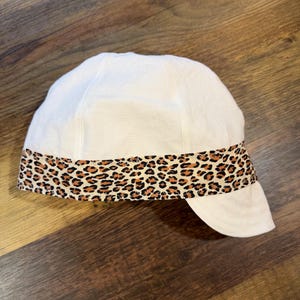 May include: A white cap featuring a horizontal band with a brown and black leopard print pattern around the base. The cap has a rounded crown and a short, curved brim extending from the front, constructed with visible stitching along the seams.