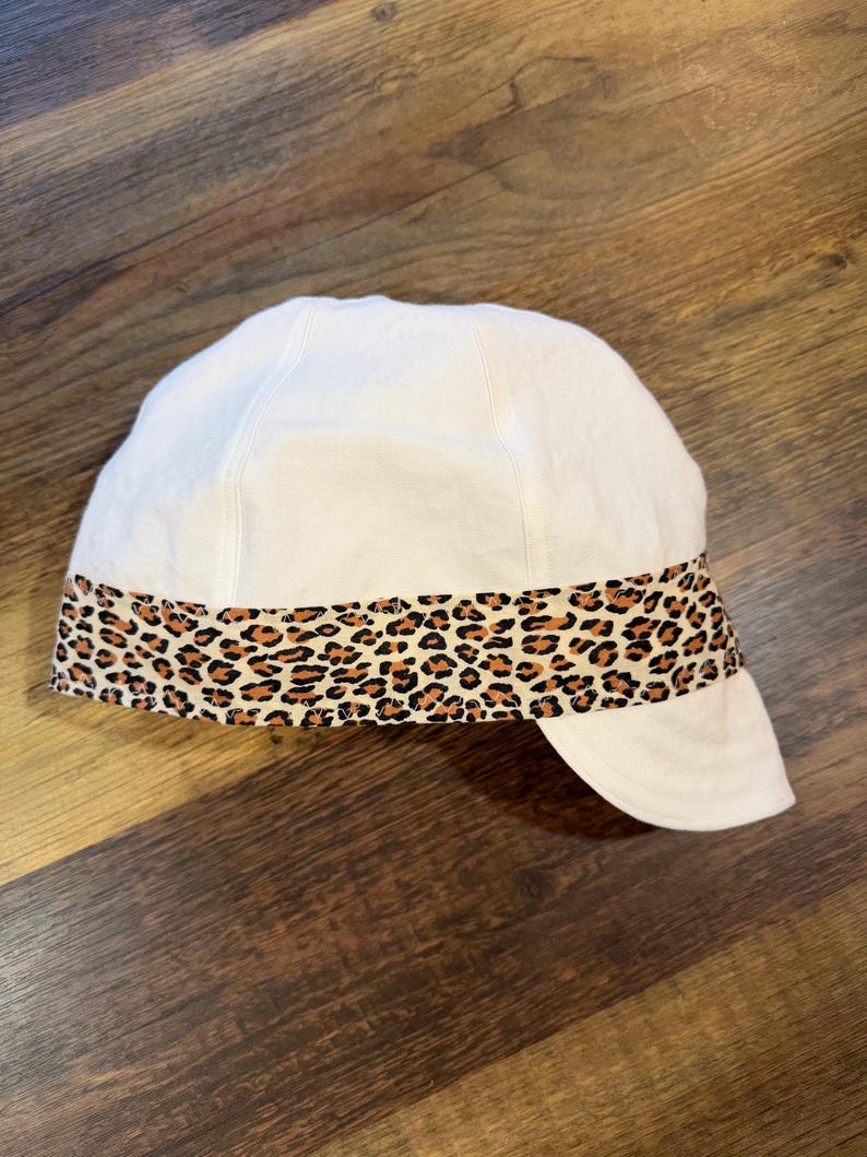 May include: A white cap featuring a horizontal band with a brown and black leopard print pattern around the base. The cap has a rounded crown and a short, curved brim extending from the front, constructed with visible stitching along the seams.