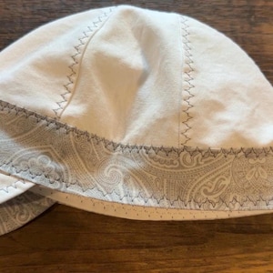 Puede incluir: Una gorra de soldador blanca con una banda de estampado de cachemir gris y una visera a juego con el mismo estampado. La gorra está construida con múltiples paneles unidos por costuras en zigzag de color gris oscuro visibles.