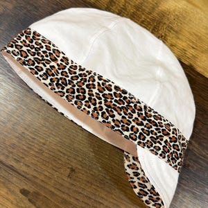 May include: A white welding cap featuring a prominent band with a brown, black, and tan leopard print pattern. The cap is constructed with multiple stitched panels and includes a curved brim section that extends from the patterned band area.