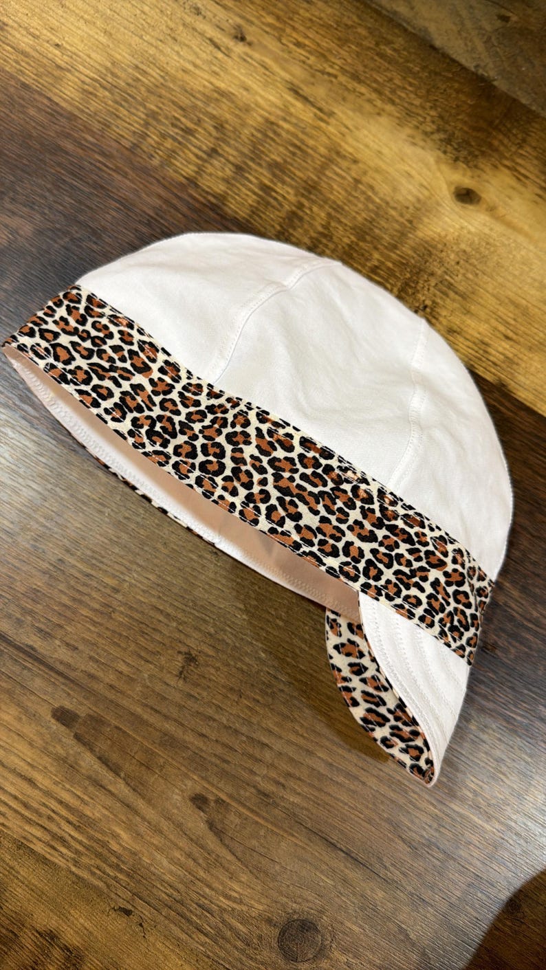 May include: A white welding cap featuring a prominent band with a brown, black, and tan leopard print pattern. The cap is constructed with multiple stitched panels and includes a curved brim section that extends from the patterned band area.