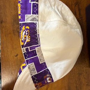 May include: A welding cap featuring a solid white crown and a wide band decorated with a purple, white, and grey geometric pattern. The band includes repeating graphics of a tiger mascot and the letters LSU, finished with white zigzag stitching.